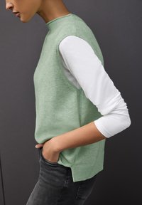 Miętowozielony bezrękawnik na białej koszuli z długim rękawem, z okrągłym dekoltem i prążkowanym wykończeniem, w zestawie z ciemnoszarymi dżinsami.