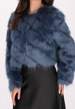 Frau trägt eine kurze blaue Kunstpelzjacke kombiniert mit einem schwarzen Satinsrock, zeigt den Oberkörper und die Hände.