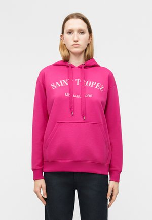 Moteris dėvi ryškiai rausvą džemperį su užrašu „Saint Tropez Michael Kors“, stovi atsipalaidavusiomis rankomis šalia kūno, priešais šviesią vientisą fono sieną.