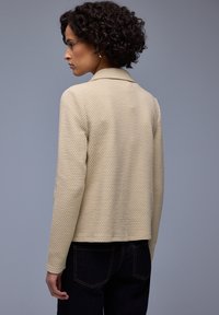 Veste beige texturée avec un col à revers et des manches longues, présentant une coupe carrée. Associée à un jean en denim bleu foncé.