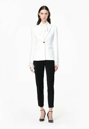 Blazer blanco con puños con volantes, cierre de un solo botón y dos bolsillos frontales, combinado con pantalones negros a medida y tacones con tiras.