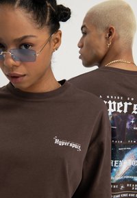 Deux personnes portant des t-shirts graphiques "Hyperspace" de couleur brun foncé, l'une faisant face à l'avant avec des lunettes bleues, l'autre montrant un design spatial détaillé dans le dos.