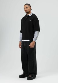 Zwarte oversized poloshirt gedragen over een grijs shirt met lange mouwen, gecombineerd met zwarte jeans met wijde pijpen en zwarte schoenen met roze veters.