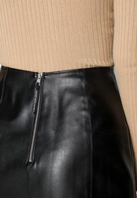 Zwarte leren rok met een hoge taille en een zichtbare metalen rits. De tailleband heeft een logo-tag. Ribbenbeige top gedeeltelijk zichtbaar.