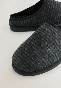 Zwarte en grijze wollen slippers met een gestructureerd oppervlak, ronde neus en zwarte rubberen zool. Rand afgewerkt met zwart stof.