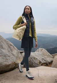 Schwarze isolierte Weste, olivgrünes Langarmshirt, schwarzer Rock und graue Wanderschuhe. Trägt eine hellbeige Tasche mit strukturiertem Muster.