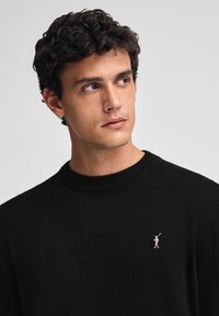 Svart stickad tröja med rundad halsringning och subtila ribbade detaljer. Har en liten, ljusrosa logotyp av en golfspelare på bröstet.