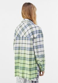 Camicia oversize a scacchi verde e bianca con dettagli blu e neri. Tessuto morbido, maniche lunghe e orlo leggermente sfilacciato.
