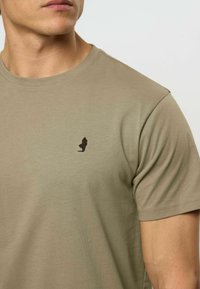 Alyvinių žalių medvilnės marškinėlių su apvalia apykakle ir trumpomis rankovėmis, su mažuoju juodu logotipu ant krūtinės ir lygiu paviršiumi.