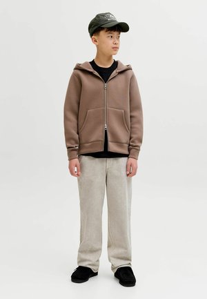 Ung person som bär en brun zip-up hoodie, svart keps, ljusbeige byxor och svarta skor, tittar åt höger mot en enfärgad bakgrund.