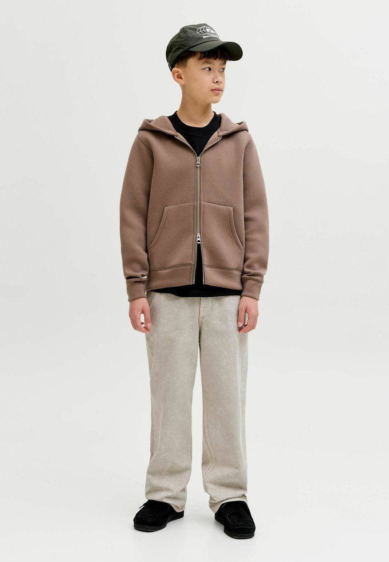 Jeune personne portant un sweat à capuche marron zippé, une casquette noire, un pantalon beige clair et des chaussures noires, regardant vers la droite sur un fond uni.