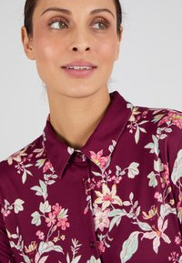 Blusa floral color burdeos con cuello, que presenta patrones florales en rosa y verde, cierre de botones y textura suave.