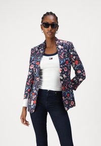 Blazer con estampado floral sobre fondo azul marino, ajuste entallado, solapas estructuradas y mangas largas; combinado con una camiseta sin mangas blanca y jeans oscuros.