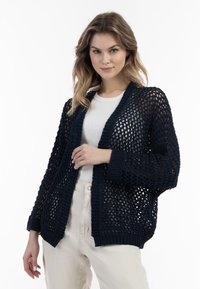 Cardigan in maglione blu navy con un design a trama aperta, maniche ampie e polsini a coste. Indossato sopra una maglietta bianca aderente e pantaloni beige.