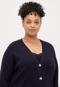 Cardigã azul-marinho feito de um material suave, com um decote em V profundo, corte relaxado e botões decorativos prateados em forma de flor ao centro.