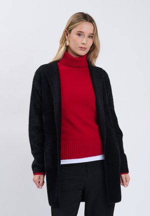 Jonge vrouw met gouden oorbellen, een rode coltrui, een zwarte cardigan en een zwarte broek, staand tegen een effen achtergrond.