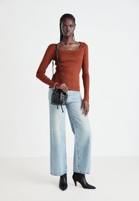 Vrouw in een roestkleurig longsleeve shirt, lichtblauwe wijde jeans, zwarte hakken en een zwarte schoudertas, staande voor een effen achtergrond.