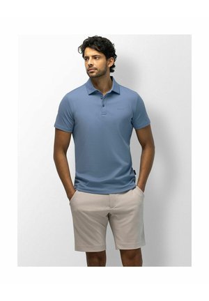 Man with dark curly hair wearing a slate blue polo shirt and beige shorts standing with hands in pockets looking left.