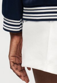Main reposant à côté d’un short blanc texturé, bord-côte blanc rayé visible au poignet et à la taille sur un pull bleu marine.
