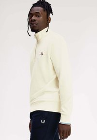 Felpa crema con zip e logo discreto sul petto, abbinata a pantaloni blu navy con logo ricamato e polsini a righe.