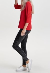 Haut rouge à manches trois-quarts associé à un jean skinny noir et des baskets blanches. Le jean présente une texture délavée, créant un look décontracté.