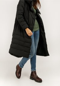 Abrigo acolchado negro con capucha, con paneles acolchados, usado sobre un suéter verde y jeans azules. Botas marrones hasta el tobillo con suelas gruesas.
