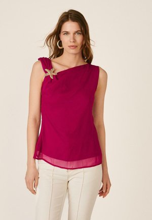 Mujer con top sin mangas magenta con broche de estrella de mar en el hombro izquierdo y pantalones blancos, de pie frente a un fondo beige.