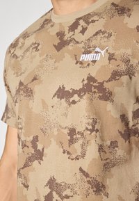 T-shirt à manches courtes de couleur beige avec un motif camouflage marron et un logo "PUMA" brodé en blanc sur la poitrine. Tissu en coton doux.