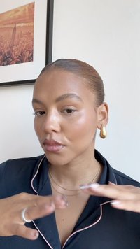 Goldene Tränenohrringe, schichtweise zarte Halsketten, dunkle Bluse mit rosa Paspel; glatte Haut mit natürlichem Make-up, Hände, die gestikulieren.