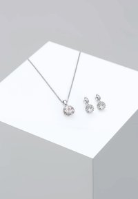 Sølv halskæde med en rund vedhæng, der har en klar sten omgivet af mindre sten; matchende studs øreringe med lignende design.