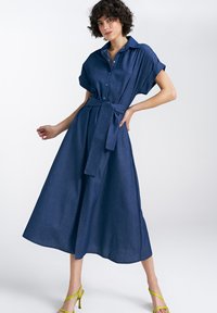 Sukienka koszulowa z denimu w ciemnoniebieskim kolorze, z krótkimi rękawami, kołnierzykiem, talia podkreśloną paskiem i zwiewną spódnicą. Połączona z jasnoniebieskimi sandałami na obcasie.