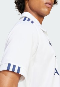 Hvid polo skjorte med krave, som har marineblå stribede detaljer på ærmerne og en glat, åndbar stof tekstur.