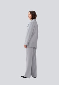 Costum pe nuanța gri deschis, cu sacou cu un singur nasture și pantaloni cu picioare ample. Textură netedă, rever tăiat și detalii minime.
