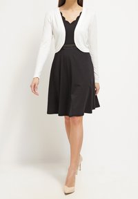 Woman wearing black scalloped neckline dress with sheer waist belt, white cropped cardigan, and beige high heels, walking forward.