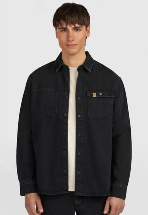 Junger Mann trägt ein aufgeknöpftes schwarzes Jeanshemd über einem weißen Hemd und steht vor einem einfarbigen hellgrauen Hintergrund.