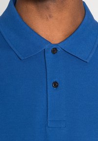 Modrá polokošile se strukturovanou tkaninou, s klasickým límcem a třemi černými knoflíky u výstřihu.