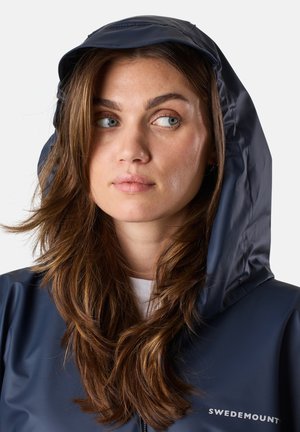 Navy waterproof jacket with a hood, featuring a smooth texture and logo on the chest. Fabric drapes naturally around the face.