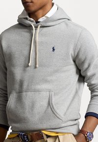 Sudadera gris con un bolsillo canguro, material de algodón, cordones crema y un detalle del logo en color marino. Presenta puños y dobladillo acanalados en color marino.
