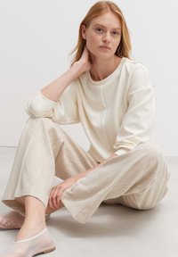 Cremefarbener Sweatshirt mit Rundhals und Bündchenärmeln, kombiniert mit hellbeigen, strukturierten, weit geschnittenen Hosen und transparenten Slippern.