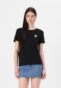 Crna majica od mekanog pamuka, s zlatnim logotipom srca na lijevoj strani prsiju. Model je kombinirao s traper suknjom, pozirajući protiv bijele pozadine.