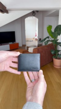 Juoda odinė piniginė su lygiu paviršiumi, dvigubo sulankstymo dizainas, matomos siūlės ir įspaustas logotipas. Laikoma rankoje šviesioje aplinkoje.