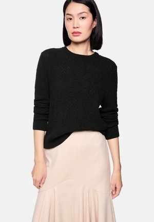 Frau mit glattem schwarzen Haar, die einen strukturierten schwarzen Pullover und einen hellbeigen Rock trägt, steht vor einem schlichten hellen Hintergrund.