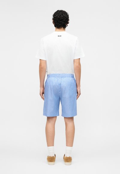 Άνδρας με σγουρά μαλλιά, φορώντας λευκό t-shirt MSGM, μπλε και λευκές ριγέ βερμούδες, λευκές κάλτσες και καφέ παπούτσια, στέκεται πλάτη.