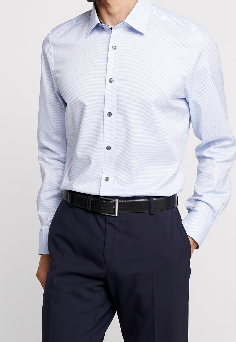Homme portant une chemise bleu clair à manches longues boutonnée, rentrée dans un pantalon bleu marine foncé avec une ceinture en cuir noire. Mains dans les poches.