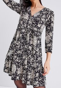 Robe noire avec des manches trois-quarts, un col en V et un ourlet volanté, présentant un motif floral blanc. Fabriquée en tissu léger.