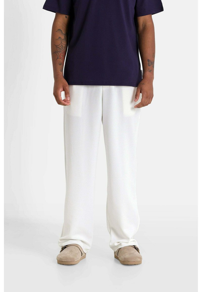 Witte wijde broek met een gladde textuur en subtiele glans, gecombineerd met een donkerpaarse oversized T-shirt en lichtbruine instap schoenen.