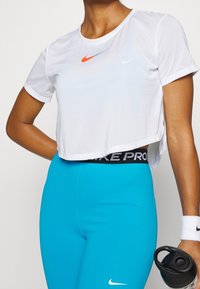 Persona vistiendo un top corto blanco y leggings de cintura alta azul brillante con logotipos de Nike, sosteniendo una botella de agua negra, con una pulsera blanca en la muñeca derecha.