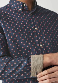 Man past, beige manchetknopen aan op een marineblauw overhemd met een rood-wit bloemmotief en parelknoopjes.