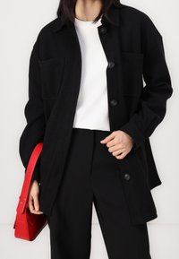 Svart överdimensionerad jacka i texturerad tyg, med stora framfickor och svarta knappar. Kombineras med en vit topp och svarta byxor, håller en röd väska.