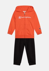 Sudadera naranja con cremallera y bolsillos frontales, adornada con el logotipo blanco de "Champion"; combinada con pantalones de chándal negros con puños acanalados. Ropa deportiva casual.